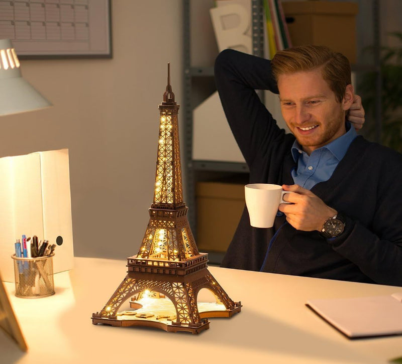 Man holding a cup in front of a lit Eiffel Tower lamp on a desk.