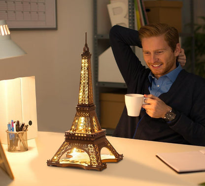 Man holding a cup in front of a lit Eiffel Tower lamp on a desk.
